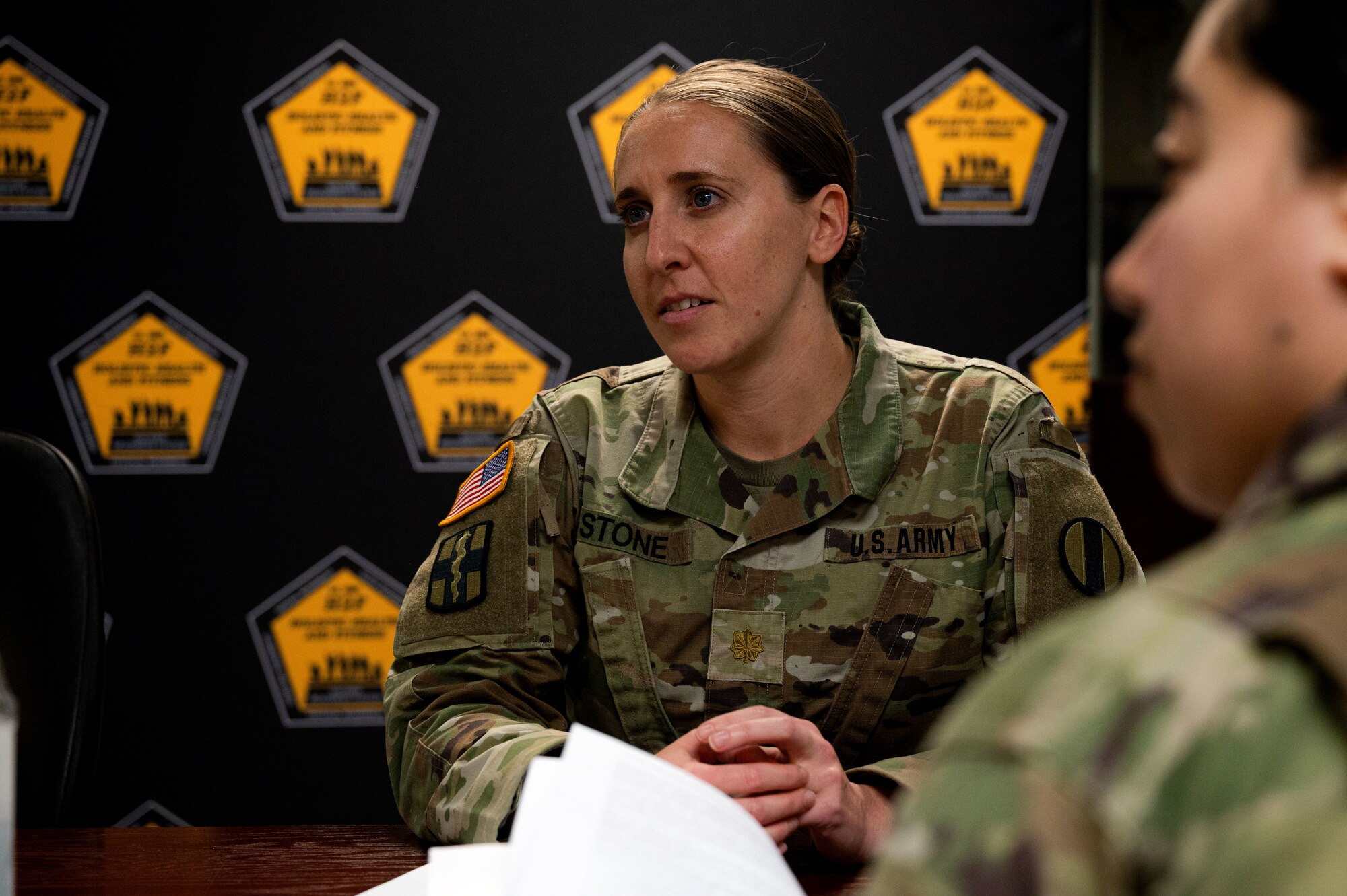 A woman in an Army uniform sits at a conference table with her hands clasped; a second woman in an Army uniform sits to the left of the first woman.