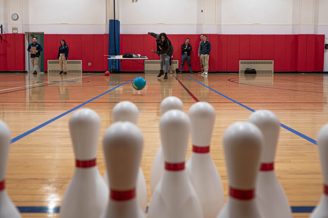 An airman rolls a frozen turkey wrapped in blue plastic down a lane toward white pins with red stripes at a bowling alley.
