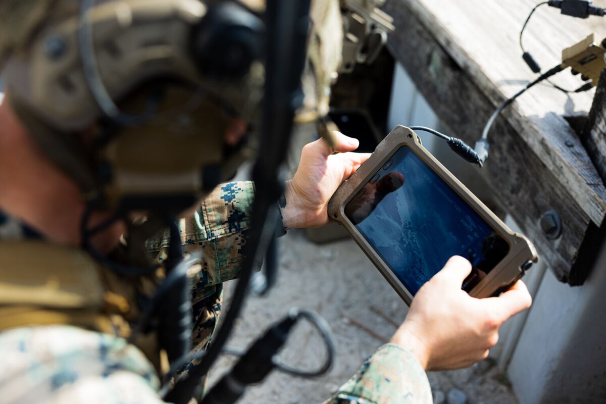 A service member wearing combat equipment looks at a handheld screen while in the field.