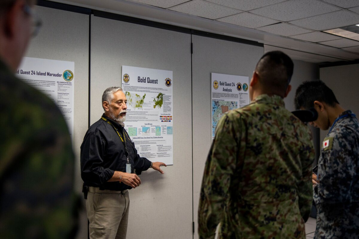 A man wearing civilian attire points to an information poster while facing foreign service members in uniform.