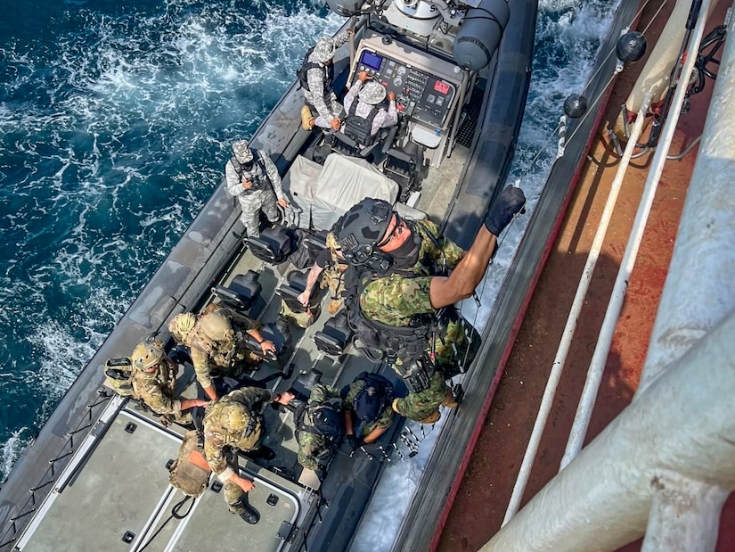 A special forces service member climbs a rope next to a ship at sea while a group of special forces remain on a boat below.