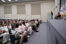 Soldier on stage behind podium speaks to audience in auditorium seats.