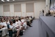 Soldier on stage behind podium speaks to audience in auditorium seats.