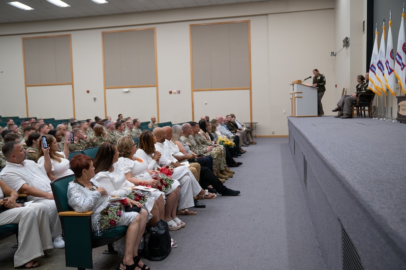 Soldier on stage behind podium speaks to audience in auditorium seats.