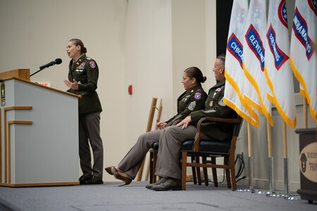 Soldier at podium speaks while looking back at two other soldiers on a stage surrounded by Army Recruiting Flags.