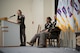 Soldier at podium speaks while looking back at two other soldiers on a stage surrounded by Army Recruiting Flags.