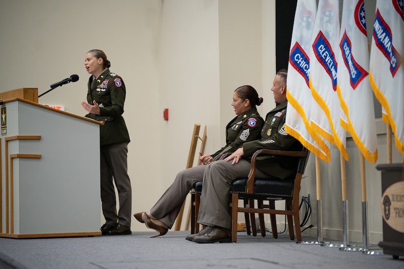 Soldier at podium speaks while looking back at two other soldiers on a stage surrounded by Army Recruiting Flags.