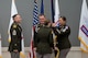 Female Soldier in dress Army uniform passes a guidon to commanding soldier while two other soldiers look on.