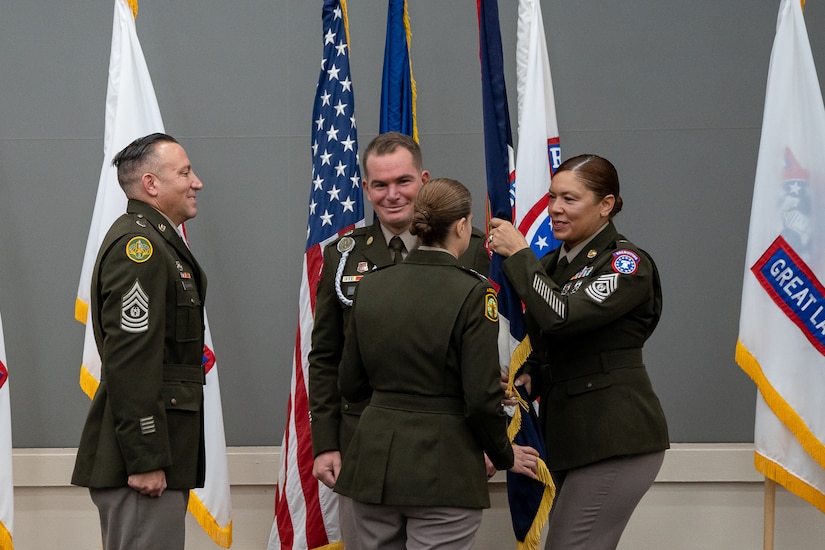 Female Soldier in dress Army uniform passes a guidon to commanding soldier while two other soldiers look on.