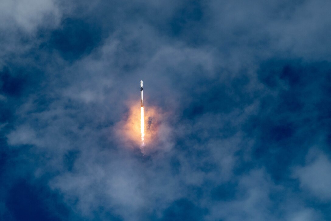 A rocket leaves a small fiery trail as it launches into a partially cloudy deep blue sky.