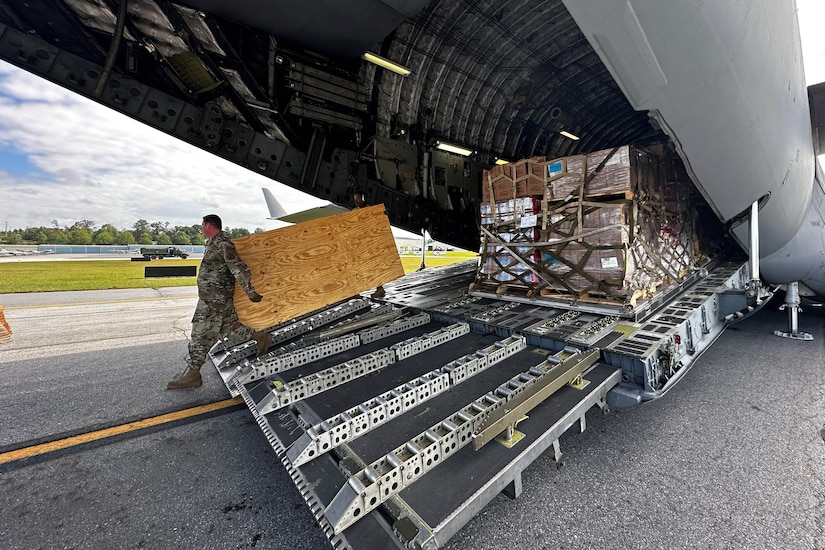 An airman stands in front of a large wooden box being unloaded from the back of an aircraft. A pallet of supplies sits on the loading platform in the background.