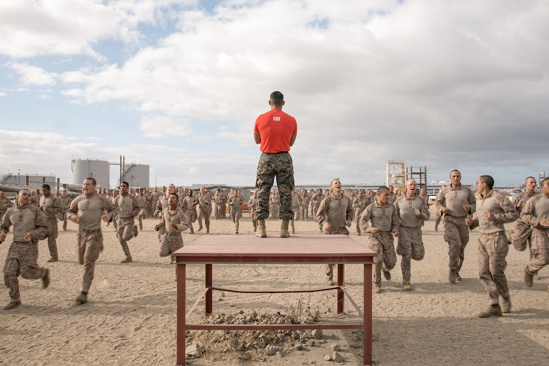 A drill instructor stands on a riser facing a large group of Marine Corps recruits as they run in a desert area.