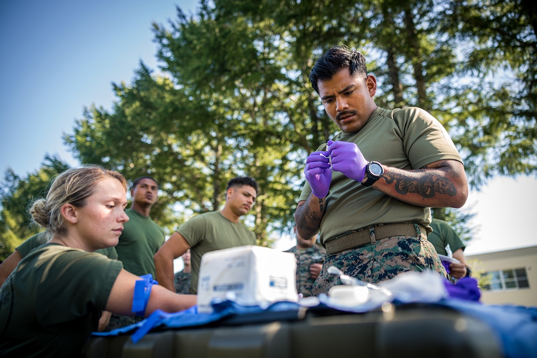 A sailor wearing gloves prepares an IV needle outdoors as another seated sailor holds out their arm. Others watch in the background.