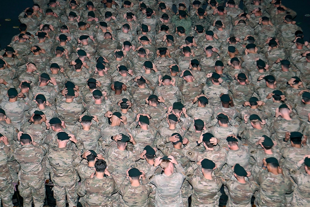 Dozens of soldiers put on green berets in a dimly lit room as seen from above.