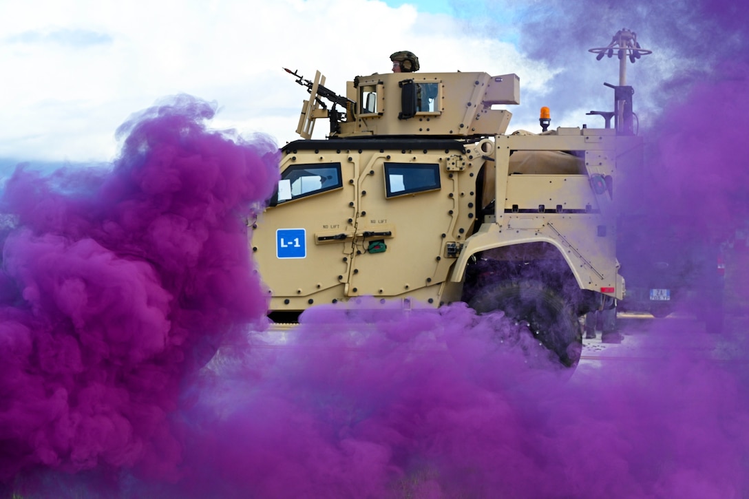 An airman aims a weapon on top of a partially visible armored vehicle as clouds of purple smoke cover the foreground.