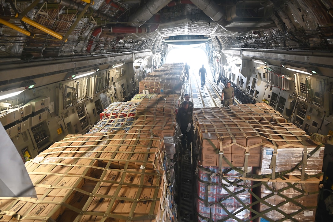 Guardsmen walk around pallets of cargo aboard an aircraft as light shines in through the open doorway.