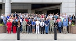 A large group of personnel stand in front of a concrete and glass building outside.