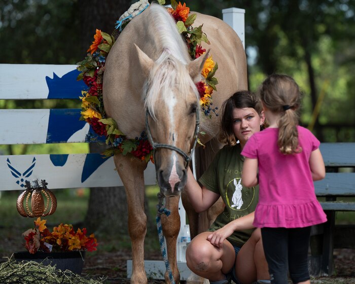 A  horse stands next to its owner as the owner kneels next to her horse and talks to a young girl.