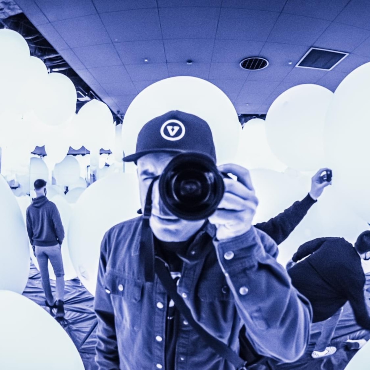 A man wearing a hat holds a camera to his face inside a room.