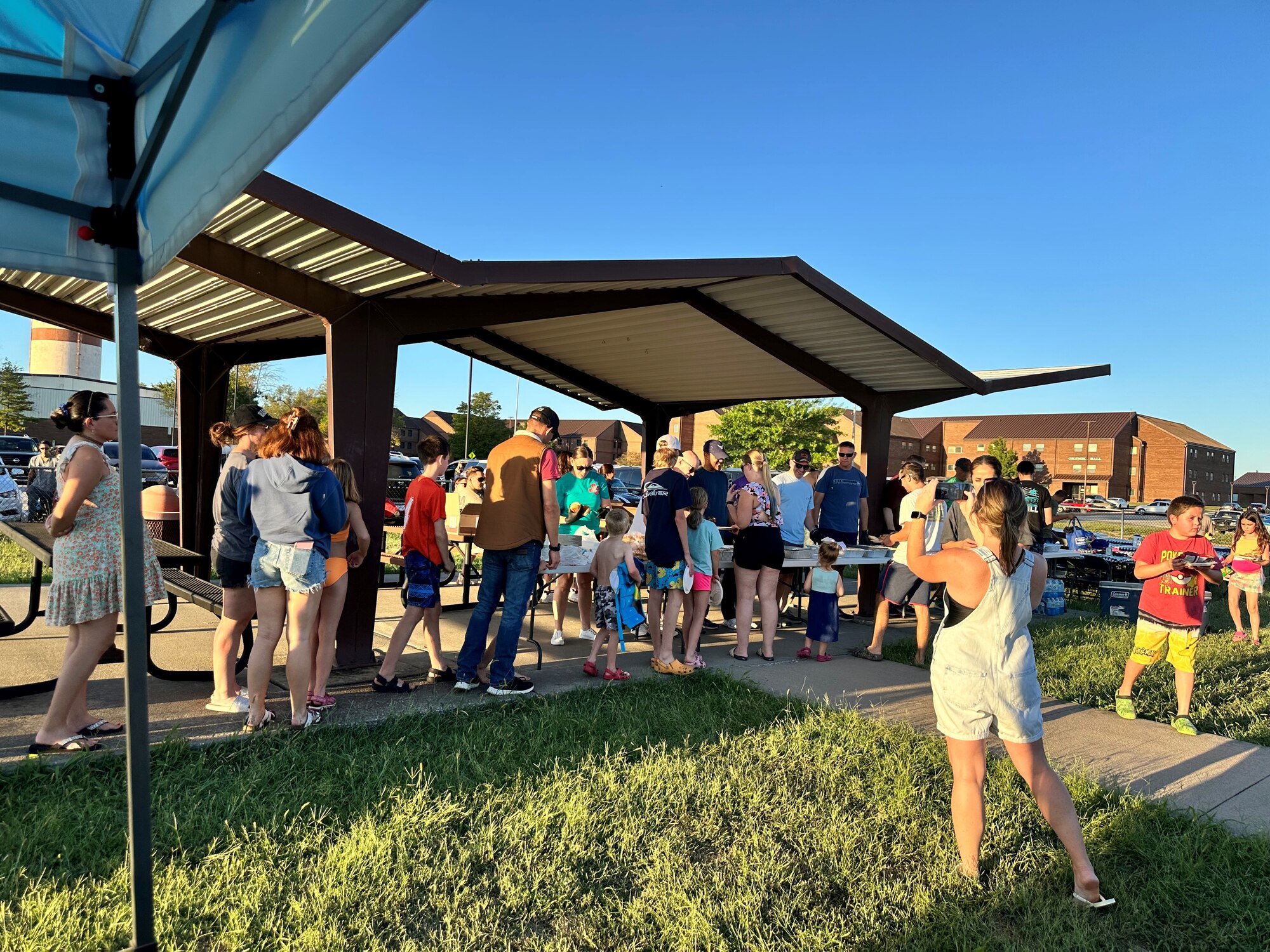 People stand together outside at an event, most people in the photo are standing in line for food at this outdoor picnic.