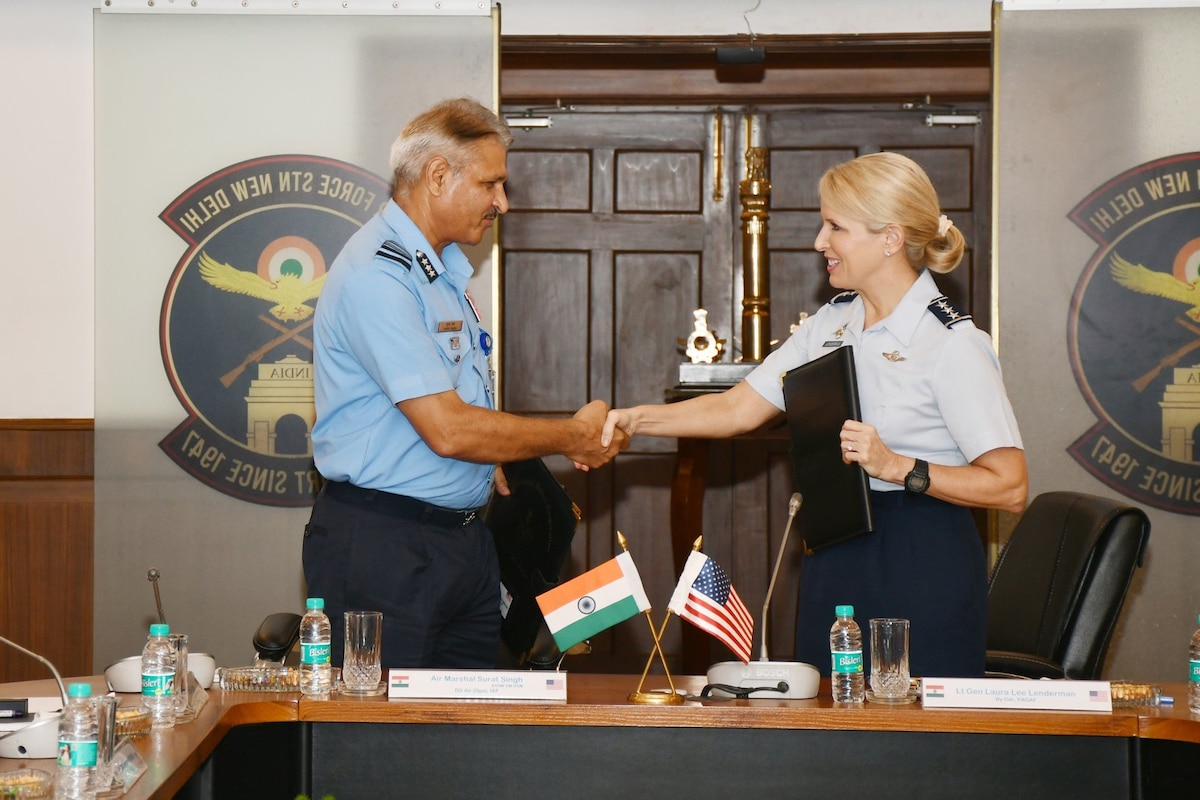 A woman in formal military attire shakes hands with a man in formal military attire in a military conference room.