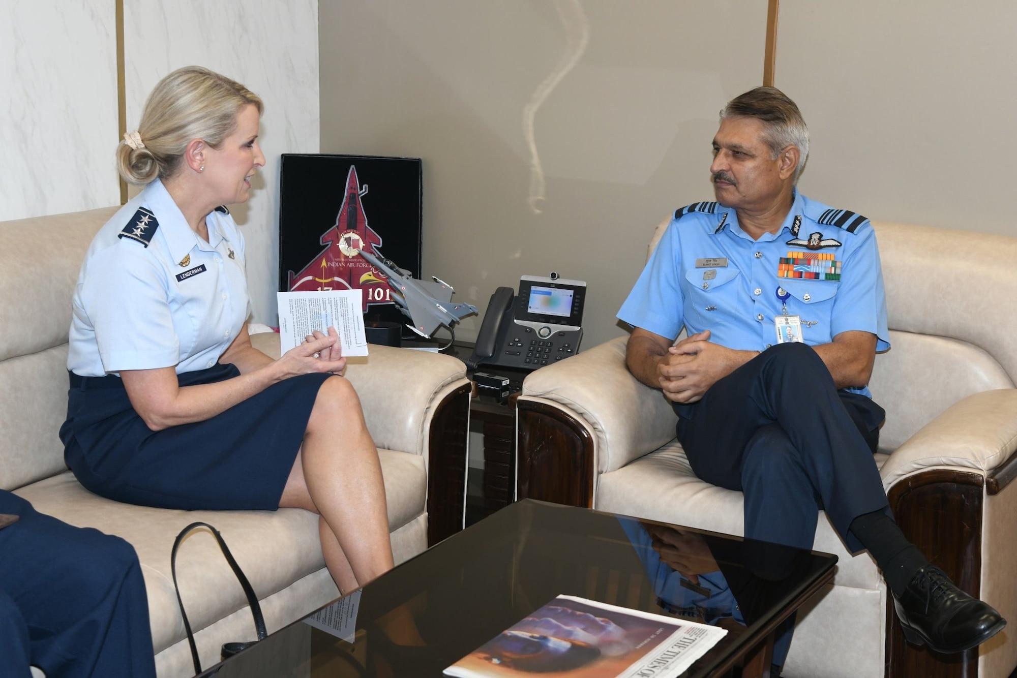 A woman in military formal attire sits on a leather couch talking to an older man in formal military attire sitting on a leather chair as they talk.
