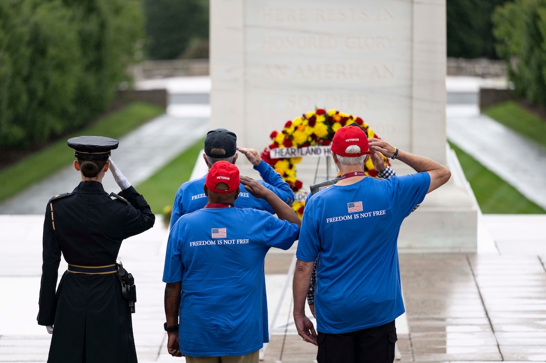 Four civilians and a soldier with their backs to the camera salute a memorial adorned by a large wreath.