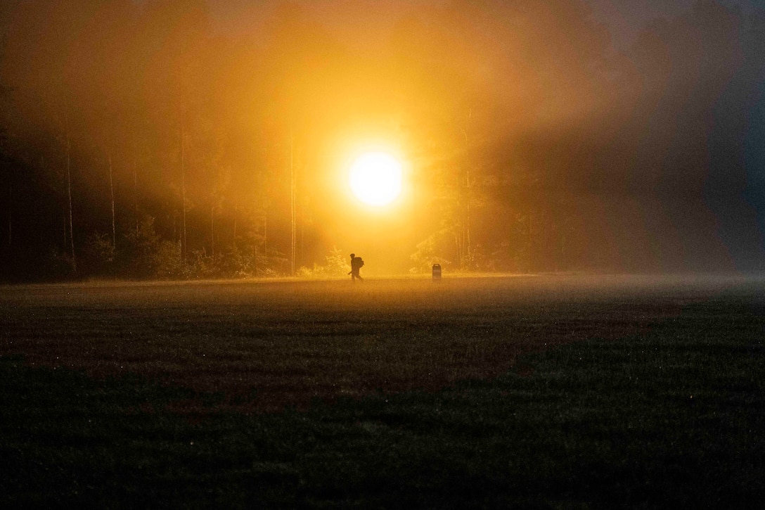 A soldier in silhouette walks across a large field at night lit up by a very bright spotlight.