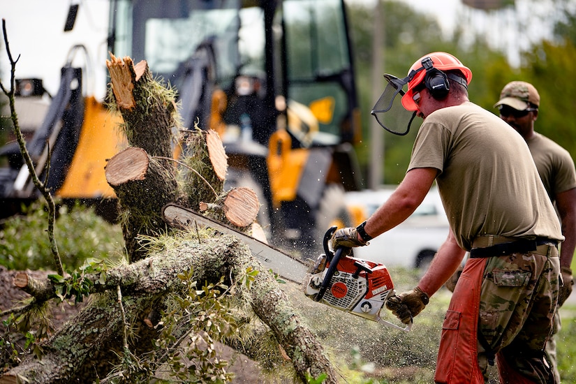 An airman wearing outdoor gear cuts a tree stump with a chainsaw during a cloudy day with a yellow tractor in the background.