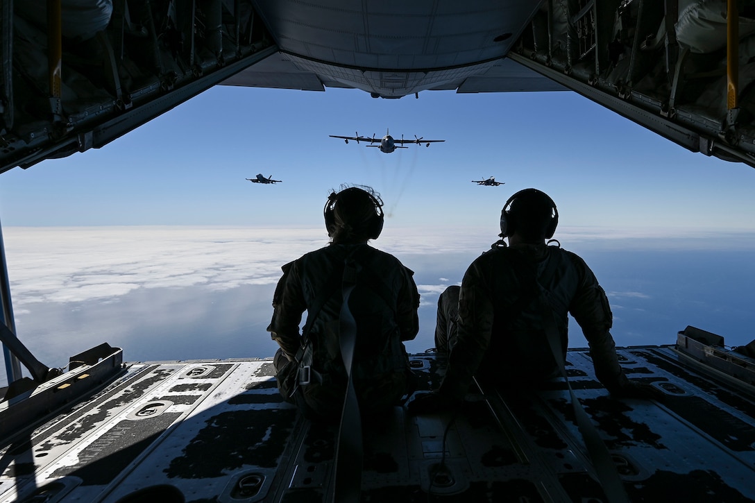 Two airman sit in the open cargo hold of an aircraft while watching three military aircraft fly behind them during daylight.