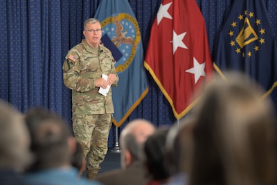 Man in camo uniform stands on a stage with the DLA and 3-star flags.