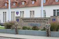 Two Soldiers in uniform pose for a photograph next to a headquarters sign.