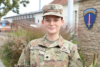 A female Soldier wearing a uniform poses for a photograph next to the US Army Europe crest.