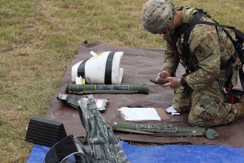 A Soldier kneels down and looks at a smart phone while working with training aides representing enemy munitions.