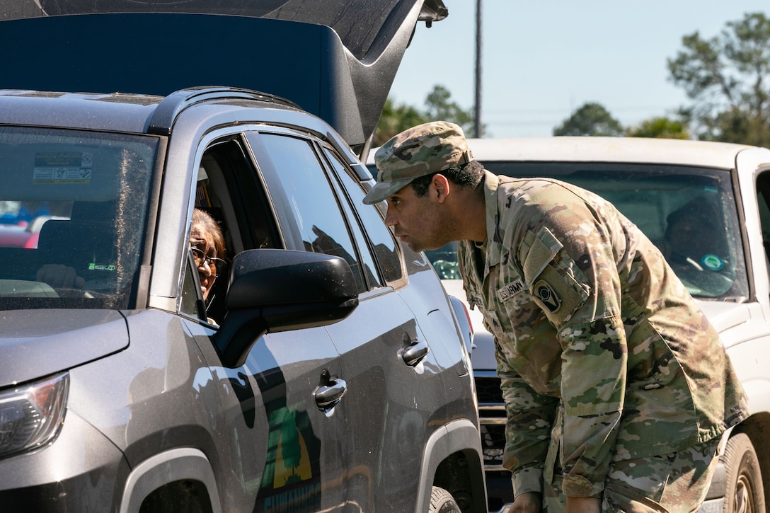 A soldier bends over to speak to a resident through their car window as other cars wait in line.