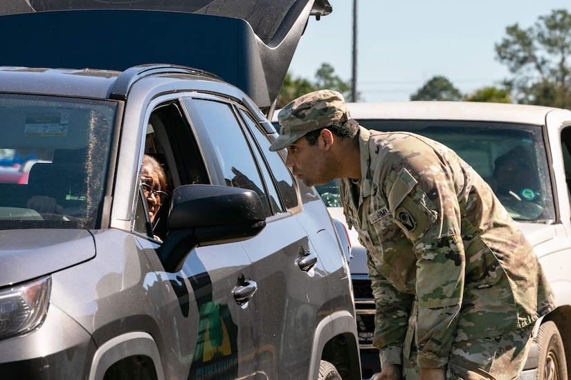 A soldier bends over to speak to a resident through their car window as other cars wait in line.