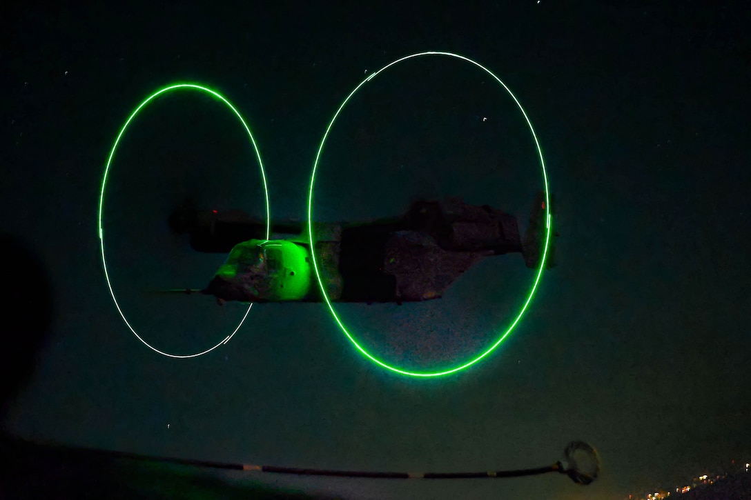 An airborne aircraft illuminated by green light approaches a fuel hose in a starry night sky.