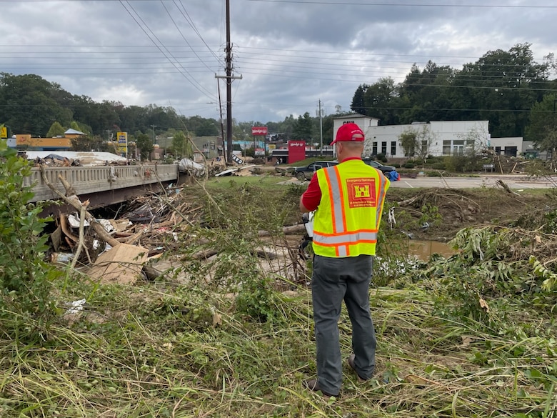 A USACE Employee in a safety vest stands on the edge of a creek bed filled with debris from recent flooding.