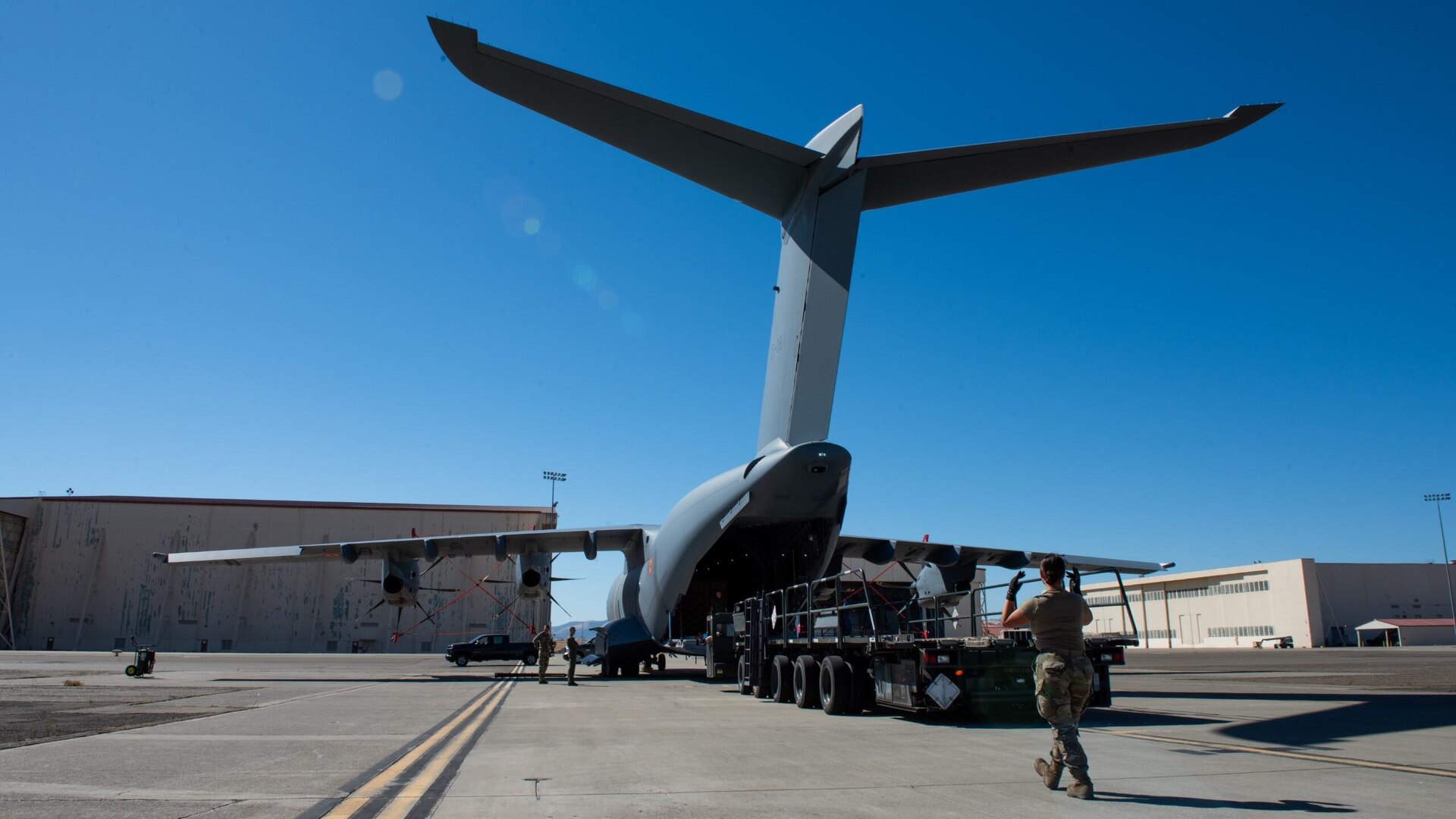 A service member walks while signaling with her hands to a cargo loader behind a military cargo plane.