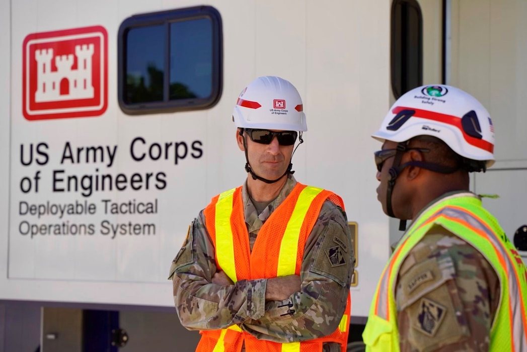 Two soldiers stand next to a large vehicle. The soldiers are wearing hard hats and safety vests over the camouflage uniforms.