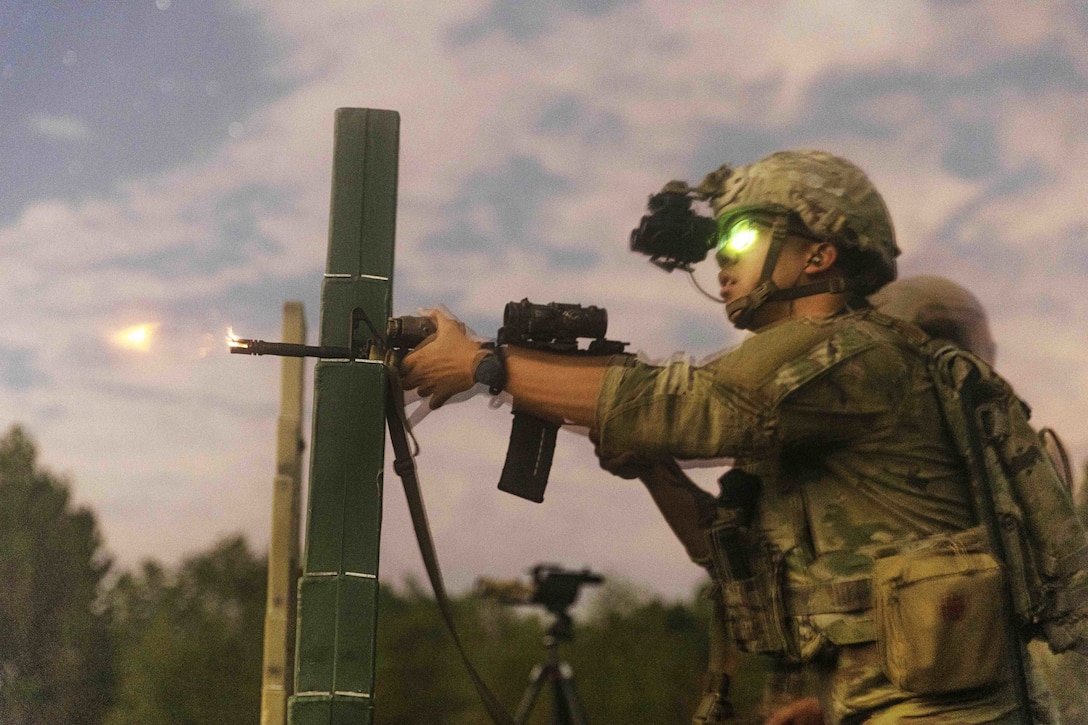 A soldier wearing a night vision device fires a weapon at a target at a shooting range in low light conditions.