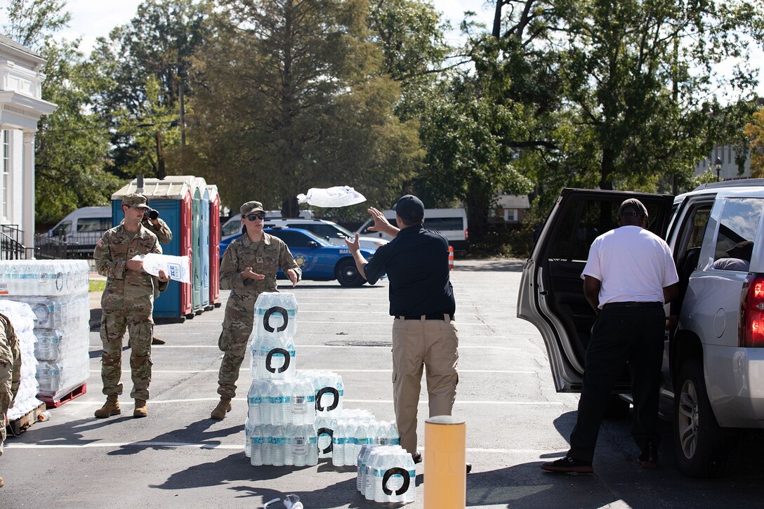 In a parking lot, a person in civilian clothes tosses a bag of ice to a soldier, while another soldier holds a bag of ice and a civilian reaches into a car.