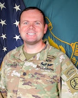 Man in U.S. Army uniform standing in front of two flags.