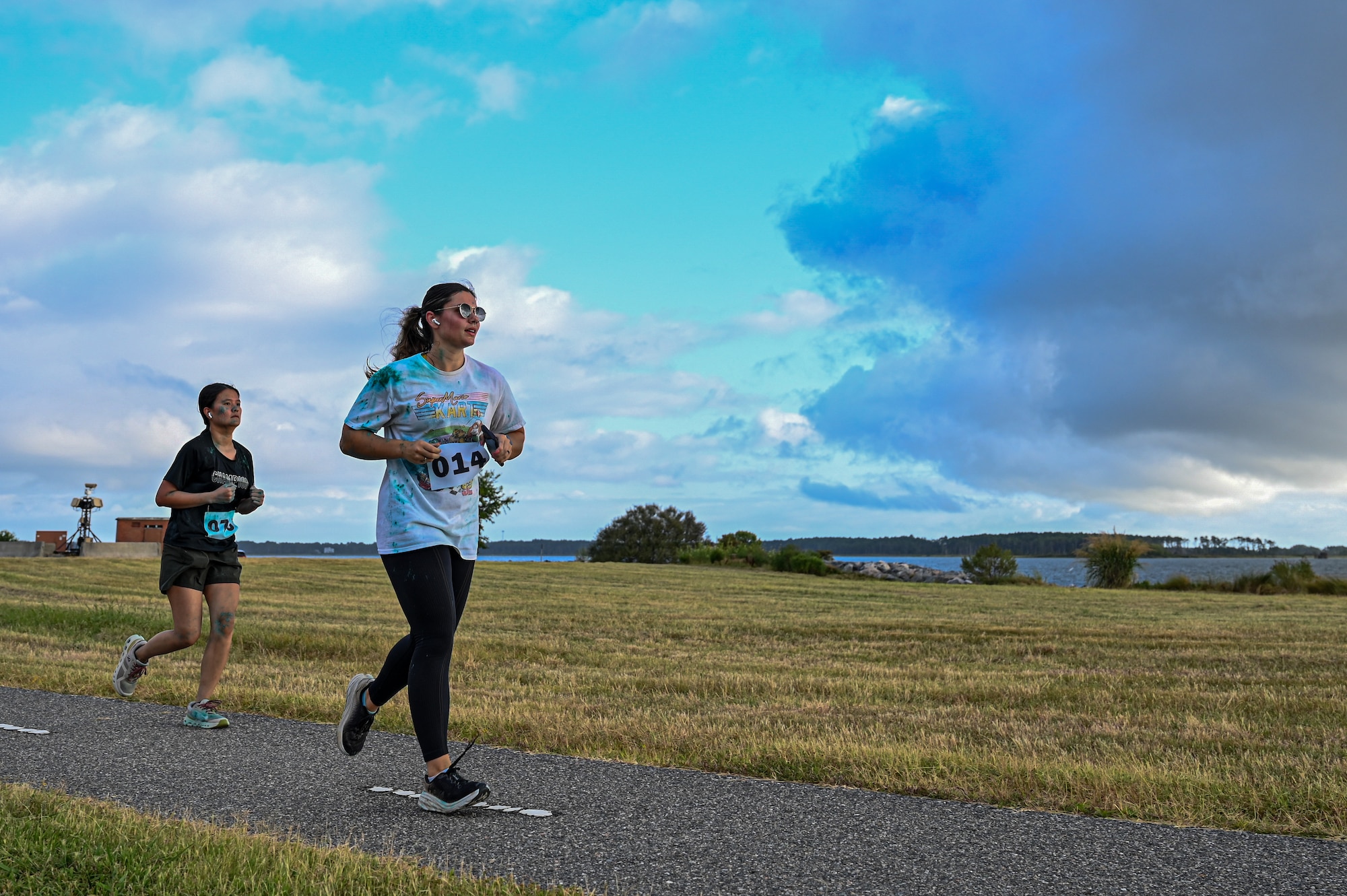 Photo of two runners running past water with clouds in the sky.