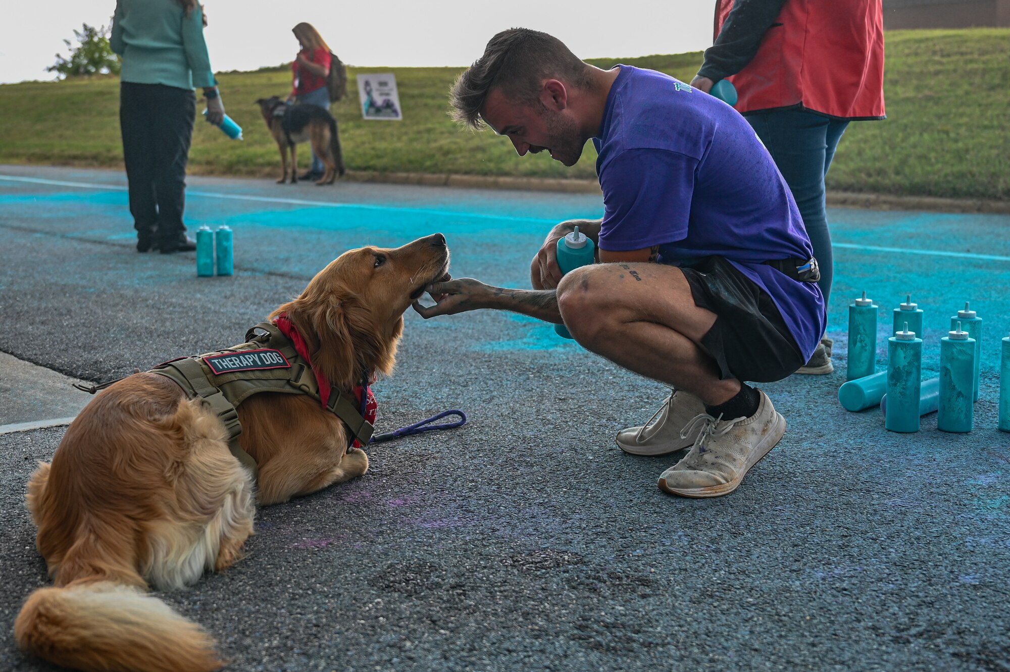 Photo of an Airman scratching the bottom of a service dogs' neck in front of location in which participants were running in a color run race.