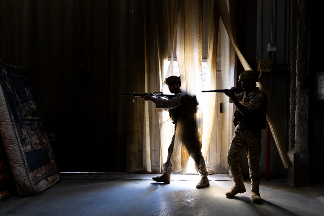 Two service members holding weapons enter a dark training room with sunlight peeking in from an entryway.