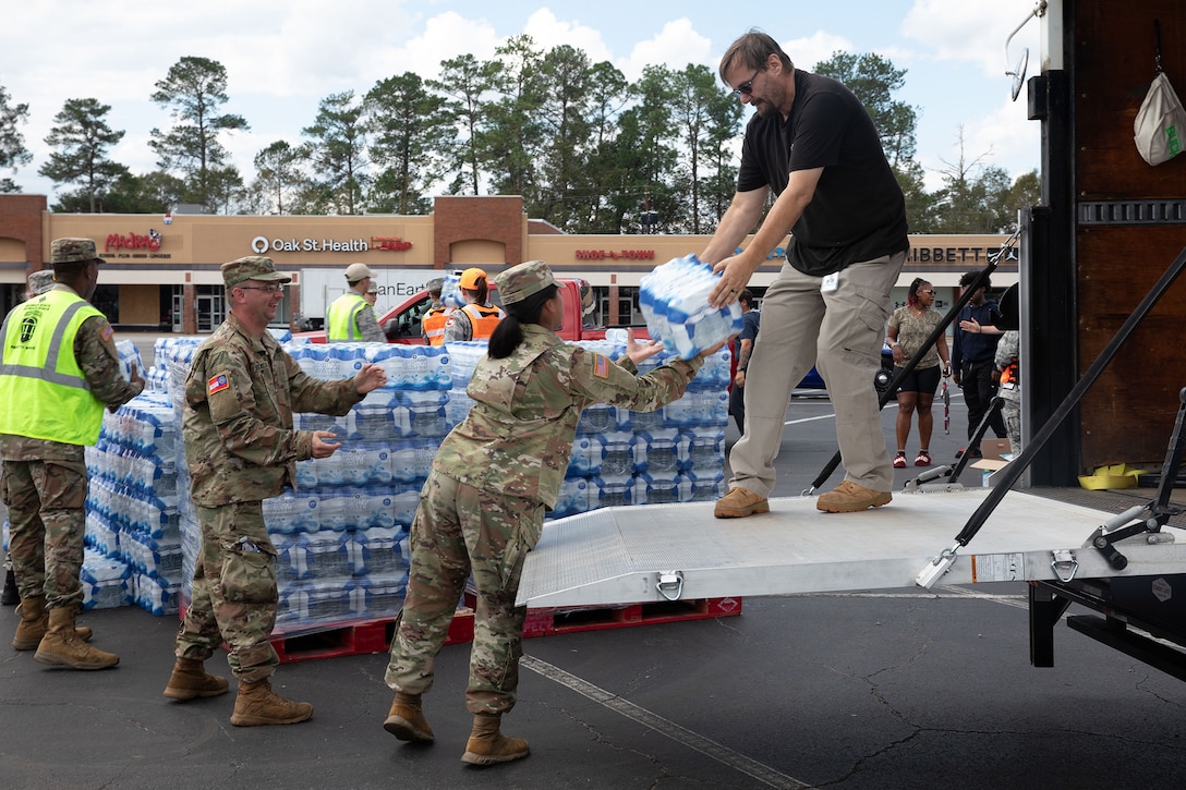 Soldiers pass packages of bottled water from a pallet to a person in civilian clothing standing on the ramp of a truck in a shopping plaza parking lot during daylight.