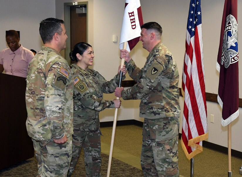 Three soldiers are standing in the photo. The soldier in the middle is handing over an "HHD" flag to another soldier.