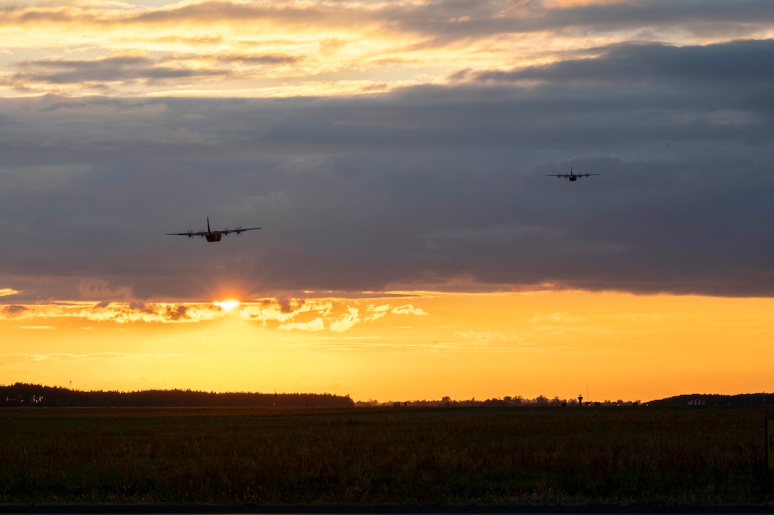 Two aircraft fly in an orangish sky as the sun peeks behind clouds in the distance.