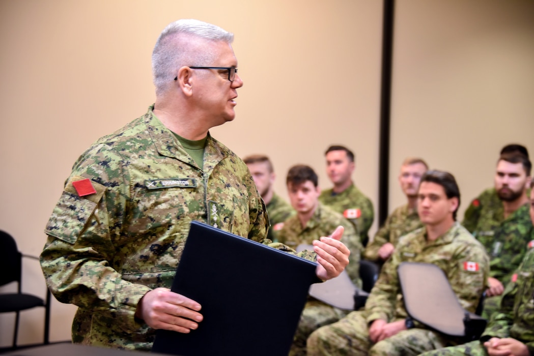 A Canadian service member address a crowd of U.S. and Canadian service members while holding an award citation.